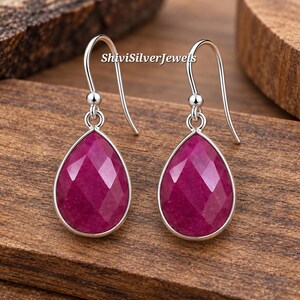 May include: A pair of silver drop earrings featuring faceted, pear-shaped magenta gemstones set in smooth silver bezels. The earrings hang from simple silver shepherd hooks. The text ShiviSilverJewels is visible in the center of the image.