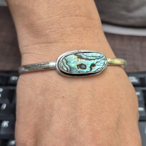 May include: Silver cuff bracelet featuring an oval abalone shell centerpiece with iridescent blues, greens, and purples. The bracelet has the text "ShivisiSilverJewels" engraved on the band.