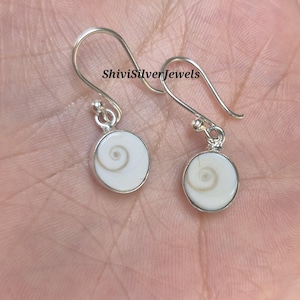 Peut inclure: Boucles d'oreilles en argent avec un centre blanc en forme de spirale. Les boucles d'oreilles ont un design en forme de crochet et sont de forme circulaire. Le texte "ShiviSilverJewels" est visible sur l'image.