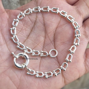 May include: A silver bracelet with a chain of U-shaped links. The bracelet has a circular clasp with a small bar closure. The bracelet is displayed on a hand. The bracelet's design is visible.