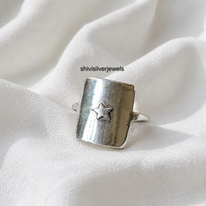 May include: A silver-toned ring featuring a large, rectangular face with a smooth, polished surface. A small, five-pointed star is embossed in the center of the rectangular plate. The text shivisilverjewels is visible above the ring.
