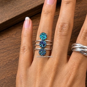 Puede incluir: Un anillo de plata con tres bandas apiladas, cada una rematada con una pequeña incrustación circular de concha de abulón con patrones iridiscentes en azul y verde. El texto ShiviSilverJewels es visible en la banda. Un anillo de plata liso se lleva en el dedo adyacente.