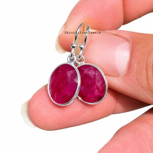 May include: A pair of oval-shaped, faceted ruby earrings set in polished silver bezels with silver hook ear wires. The deep pinkish-red gemstones feature a textured, multifaceted surface that catches the light, suspended from simple silver loops.
