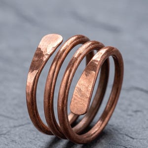 Könnte beinhalten: Ein Kupferring mit einem Spiraldesign aus drei deutlichen Windungen. Die Metalloberfläche weist eine gehämmerte Textur auf, und die beiden Enden des Drahtes sind abgeflacht und verjüngt, was diesem Schmuckstück ein umlaufendes Aussehen verleiht.
