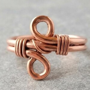Può includere: Un anello in filo di rame caratterizzato da un design centrale composto da due anelli sovrapposti. La fascia è realizzata con più fili di rame lucido, con segmenti di filo più sottili avvolti strettamente sui lati per fissare la struttura.