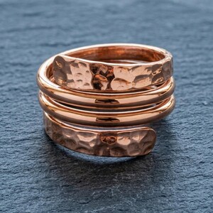 Könnte beinhalten: Ein kupferfarbener Ring mit einem mehrlagigen Spiraldesign. Die beiden äußeren Enden des Rings weisen eine texturierte, gehämmerte Oberfläche auf, während die beiden mittleren Bänder glatt und poliert sind, was einen geschichteten Glanz erzeugt.
