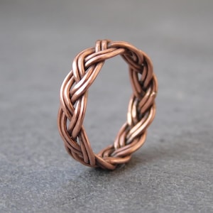 Könnte beinhalten: Ein Ring aus mehreren miteinander verflochtenen Kupferdrähten in einem durchgehenden Zopfmuster. Das Metall hat eine warme, rotbraune metallische Oberfläche mit polierter Textur und bildet einen kreisförmigen Ring mit einem komplexen Geflecht.