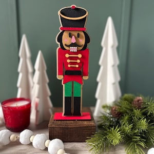 May include: A wooden Christmas nutcracker figurine with a red coat, green pants, and a black hat. The figurine stands on a wooden base, with white Christmas trees and a red candle in the background. A festive holiday decoration.