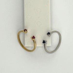 May include: Two open-hoop earrings. One is gold-toned with red gemstones, the other is silver-toned with black gemstones. The earrings are displayed on a white background.