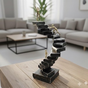 Peut inclure: Une sculpture en bronze représentant un escalier fait de livres, avec de petites figurines dorées qui montent les marches. Une figurine est au sommet, les bras levés. La sculpture est sur une table en bois.