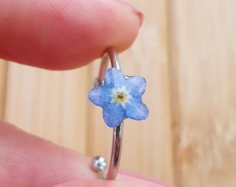 May include: A silver-colored ring featuring a delicate, light blue forget-me-not flower. The flower has a yellow center and is set on a thin, curved band. The ring is held between two fingers, showcasing the floral design.