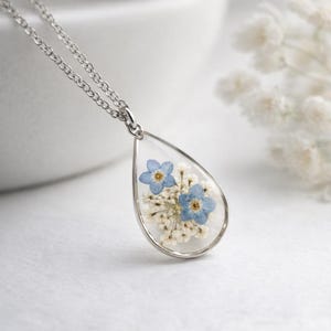 May include: A silver necklace featuring a teardrop pendant. The pendant contains blue forget-me-not flowers and tiny white flowers set in clear resin. The chain is fine, and the pendant is the focal point.