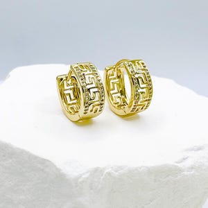 May include: A pair of gold-colored hoop earrings with a Greek key design. The earrings are small and round, with a hinged closure. The design is repeated around the entire hoop. The earrings are displayed on a white, textured surface.