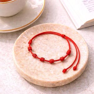 May include: A red string bracelet with multiple knots, resting on a round, beige marble coaster. A white teacup with tea and a gold rim sits nearby, along with a partially visible book.