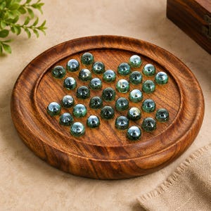 May include: A round wooden solitaire board game featuring a dark brown grain finish with a recessed circular playing area. The board is filled with numerous translucent, dark green glass marbles arranged in a cross-shaped pattern across the surface.