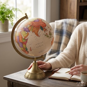 Pode incluir: Um globo de estilo vintage com um mapa creme e continentes coloridos. O globo está montado em um suporte de latão e está em uma mesa de madeira. Uma pessoa está segurando uma caneta e um caderno. O diâmetro do globo é de cerca de 30 cm.