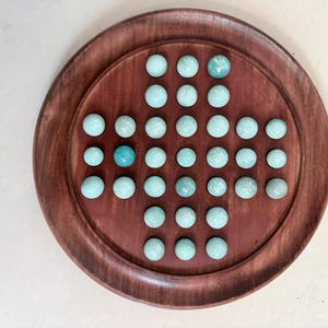 May include: A circular wooden board game featuring a cross-shaped grid of indentations. The board is filled with smooth, light blue, spherical marbles, with one marble positioned in a slightly darker shade of blue within the grid pattern.