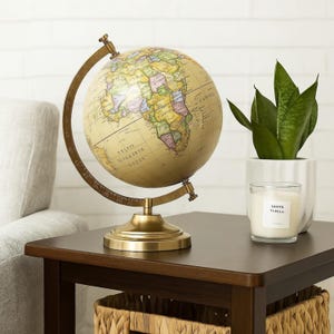 Peut inclure: Un globe de style vintage avec une carte crème et un support couleur laiton. Le globe est posé sur une table marron foncé, à côté d'une plante en pot et d'une bougie. La bougie a une étiquette blanche avec du texte noir.