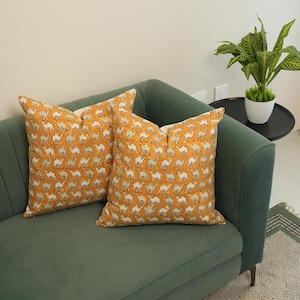 Pode incluir: Duas almofadas decorativas com um padrão de camelo sobre um fundo laranja. As almofadas são colocadas num sofá verde. O padrão de camelo é branco e cinzento. As almofadas adicionam um toque decorativo à sala de estar.