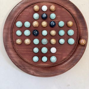 May include: A circular wooden board game featuring a cross-shaped grid of indentations filled with various glass marbles. The marbles are colored in shades of light blue, tan, white, and dark blue, arranged across the polished brown wood surface.