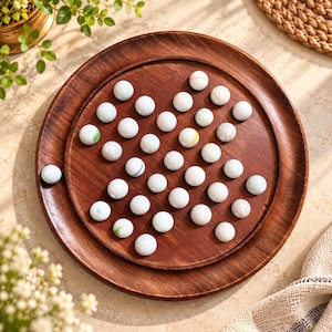 May include: A round wooden board game featuring a star-shaped pattern of indentations filled with smooth, white glass marbles. The board has a dark brown, polished finish with a raised outer rim, and one marble sits outside the board.
