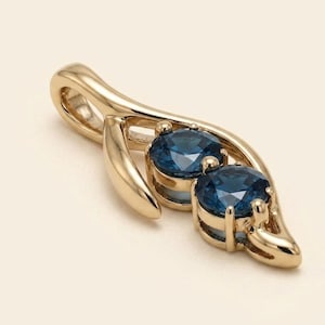 Pode incluir: Um pingente de ouro com duas pedras preciosas redondas e facetadas em azul escuro, fixadas em uma estrutura de ouro polido. O engaste de metal tem um design liso e curvo em forma de folha que se estreita em direção a uma argola superior.
