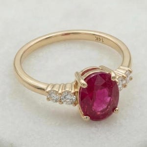 May include: A gold ring with a large oval ruby gemstone at the centre, with smaller, round, clear gemstones on either side. The ring band is polished gold. The ring is displayed on a white surface. The ring is marked '14K'.