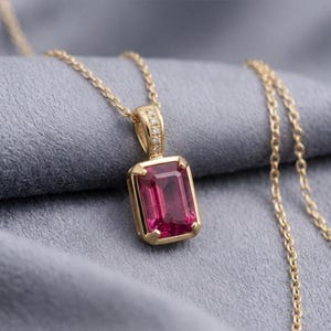 Könnte beinhalten: Eine Goldhalskette mit einem rechteckigen, im Smaragdschliff geschliffenen, tiefrosa Edelstein in einer Goldfassung. Die Öse ist mit kleinen weißen Steinen besetzt. Der Anhänger hängt an einer zarten Goldkette und wirkt sehr elegant.