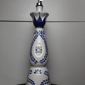 May include: A tall white ceramic bottle with a tapered neck and a flared top, decorated with hand-painted blue leaf patterns and a central floral emblem. The bottle is topped with a silver-coloured metal pourer spout with a black rubber seal.