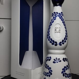May include: A white ceramic tequila bottle with blue hand-painted leaf patterns and a silver-toned bell-shaped stopper, standing next to its matching white rectangular box labelled Clase Azul Mexico Tequila Reposado with blue accents.