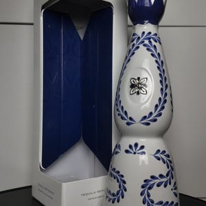 May include: A white ceramic tequila bottle with blue hand-painted leaf patterns, a silver-coloured metallic cap, and a central silver emblem. The bottle stands next to its white and blue cardboard packaging box labelled with the text Clase Azul.