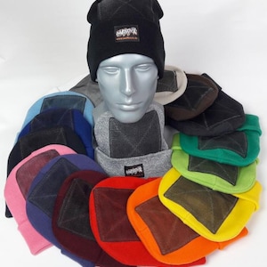 Könnte beinhalten: Eine Sammlung farbenfroher Mützen, die auf einem Schaufensterkopf ausgestellt sind. Die Mützen sind in verschiedenen Farben erhältlich, darunter Schwarz, Rosa, Blau, Grün und Orange. Jede Mütze hat ein quadratisches Mesh-Design und ein kleines Etikett mit Text. Die Mützen sind aus weichem Strickmaterial.