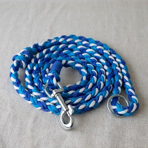 Puede incluir: Correa trenzada para perros en tonos azules y blancos. La correa está enrollada, mostrando el intrincado patrón de la cuerda entrelazada. Un mosquetón y un anillo de metal plateado están unidos para asegurar al collar.