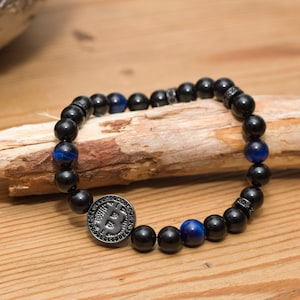 Può includere: Braccialetto di perline nere con un ciondolo con il simbolo Bitcoin. Il braccialetto presenta perline nere e blu, con un ciondolo Bitcoin color argento al centro. Il braccialetto è esposto su una superficie di legno.