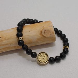 Può includere: Braccialetto di perline nere con un simbolo Bitcoin color oro e dettagli. Il braccialetto presenta perline nere opache e lucide, con distanziatori color oro. Il simbolo Bitcoin è al centro del braccialetto.
