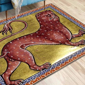 Op de afbeelding: Een rechthoekig tapijt met een middeleeuwse leeuwenillustratie in rood en bruin met witte stippen, op een gouden achtergrond. Het tapijt heeft een decoratieve rand met blauwe, rode en witte geometrische patronen. Het tapijt is waarschijnlijk van stof.