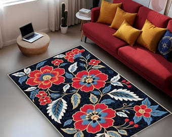 Puede incluir: Una alfombra rectangular con diseño floral. La alfombra presenta grandes flores rojas con centros azules y amarillos, rodeadas de hojas azules y blancas, sobre un fondo azul oscuro. La alfombra está colocada sobre un suelo gris cerca de un sofá rojo.