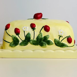 Könnte beinhalten: Eine gelbe Keramik-Butterdose mit Deckel, verziert mit roten Erdbeeren, grünen Blättern und weißen Blüten. Die Schale hat einen gewellten Rand und eine rechteckige Basis. Der Deckel hat eine einzelne Erdbeere oben.
