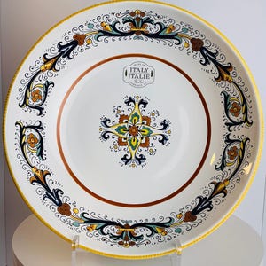 May include: A round ceramic plate with a white base, featuring a yellow rim and a central floral motif in yellow, blue, and brown. The border displays intricate scrollwork patterns. A stamp in the center reads MADE IN ITALY ITALIE.