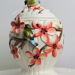 May include: A white ceramic jar with a lid featuring a three-dimensional design of brown branches, pink cherry blossoms, green and blue birds, and a small red ladybug. The jar has a vertical ribbed texture and a rounded, tapered base shape.