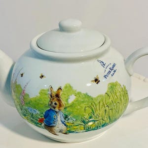 Può includere: Teiera in ceramica bianca con un design di Peter Rabbit. La teiera ha un coperchio, un beccuccio e un manico. L'illustrazione raffigura Peter Rabbit con una giacca blu, circondato da api e un paesaggio erboso. Le parole "Peter Rabbit" sono stampate sul lato.