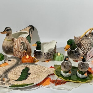 May include: A collection of ceramic duck-themed kitchenware including a large duck-shaped bowl, a smaller duck bowl, a duck-shaped platter, a duck teapot with a bamboo handle, and two small duck salt and pepper shakers on a green leaf-shaped tray.