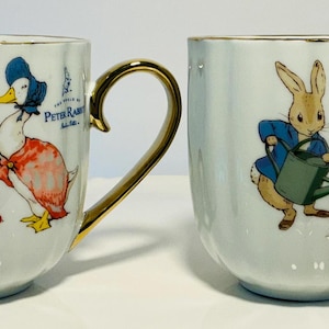 May include: Two white ceramic mugs with gold handles and rims. Each mug features a colorful illustration of Peter Rabbit and a duck. The mugs are on a white, scalloped plate.