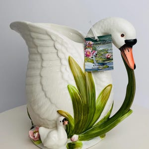 Peut inclure: Pichet en céramique blanche en forme de cygne avec une longue anse incurvée. Le pichet représente une tête et un cou de cygne, avec des roseaux verts et jaunes, un petit cygne et des fleurs roses. L'étiquette indique "Swan and Lotus".