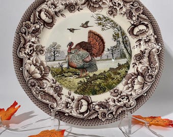 May include: A round ceramic plate featuring a central illustration of a turkey in a landscape with trees and birds. The rim is decorated with a brown and white floral pattern. The plate is displayed on a clear stand with orange maple leaves.