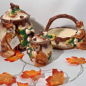 May include: A ceramic kitchen set featuring a large canister, a smaller canister, and a handled basket. Each piece is decorated with sculpted foxes, acorns, and oak leaves in shades of brown, orange, and green. A tag on the large canister reads Heritage Harvest.