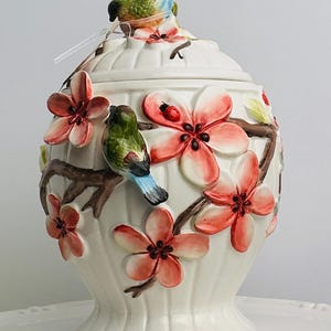 Puede incluir: Un tarro de cerámica blanca con tapa que presenta un pájaro tridimensional en la parte superior. El tarro está decorado con ramas marrones, flores de cerezo rosas, hojas verdes y una pequeña mariquita roja en relieve y pintadas a mano.