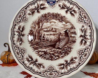 May include: A cream-colored ceramic plate featuring a brown transferware design of two pheasants in a landscape with trees and a fence. The border displays an intricate pattern of oak leaves and acorns. A Royal Stafford label is at the top.