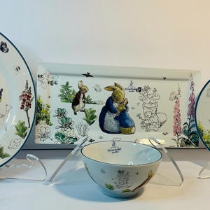 May include: A Peter Rabbit themed dinnerware set featuring plates, a bowl, and a serving tray. The white ceramic pieces have a blue rim and are decorated with illustrations of Peter Rabbit and other characters in a garden setting.