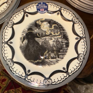 Puede incluir: Un juego de platos decorativos con una ilustración en blanco y negro de una escena espeluznante. Los platos tienen un borde festoneado con cintas negras y diseños de calabazas. El centro del plato presenta una figura con un abrigo largo frente a una casa embrujada.